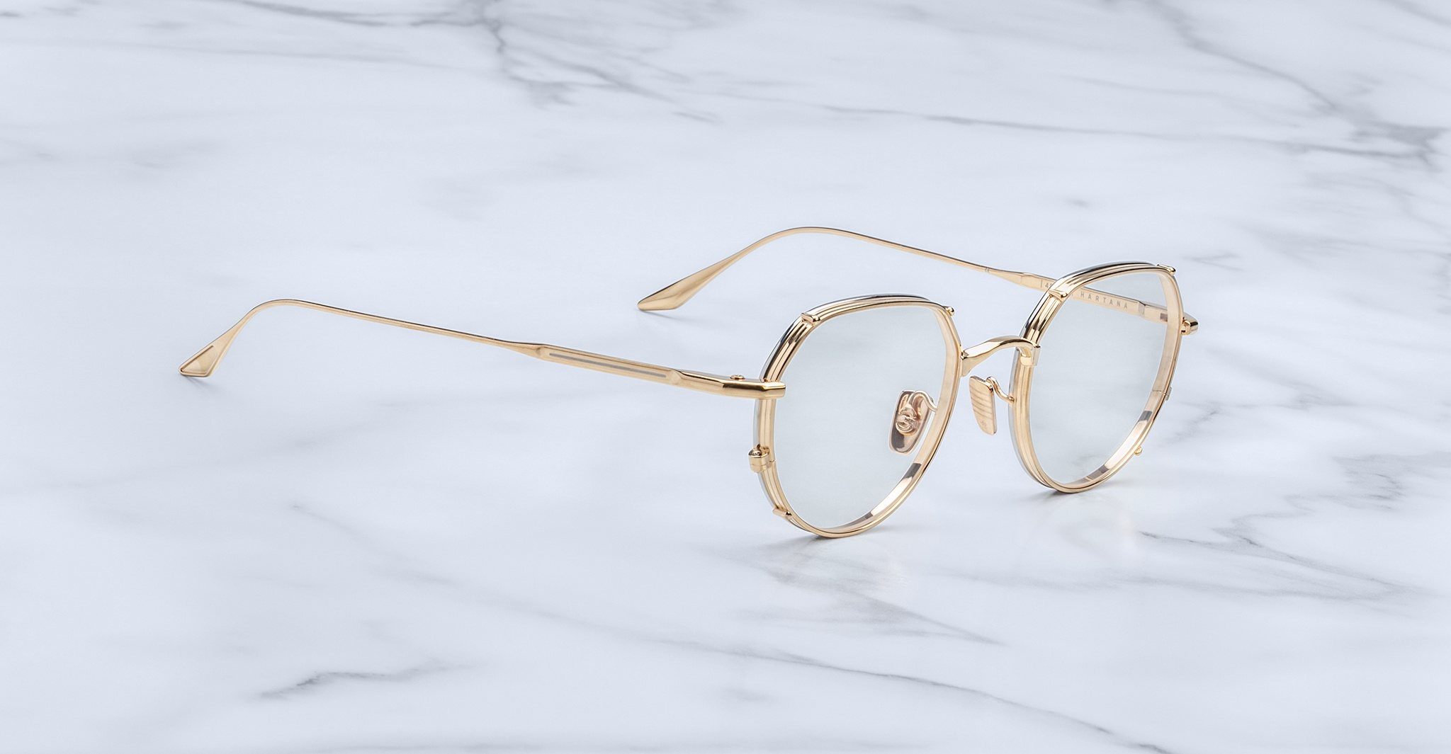 Jacques Marie Mage Hartana eyeglasses with a gold metal frame and clear lenses are displayed on a white marble surface.