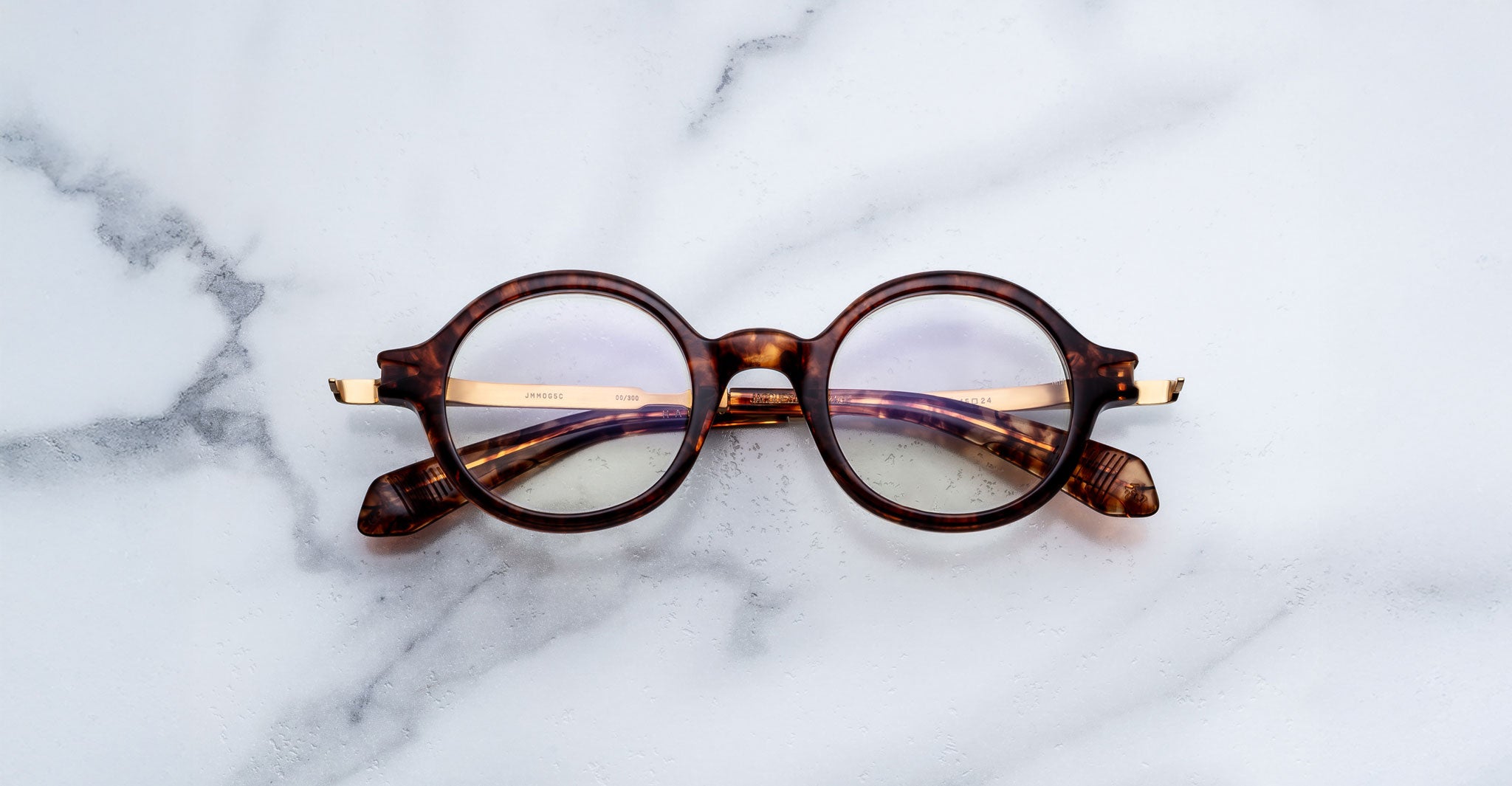 Jacques Marie Mage Hartigan in Argyle, round tortoiseshell acetate eyeglasses with a keyhole bridge, exposed metal core temples, and subtle purple anti-reflective lens coating, shown front-facing on a light marble surface.