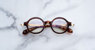 Jacques Marie Mage Hartigan in Argyle, round tortoiseshell acetate eyeglasses with a keyhole bridge, exposed metal core temples, and subtle purple anti-reflective lens coating, shown front-facing on a light marble surface.