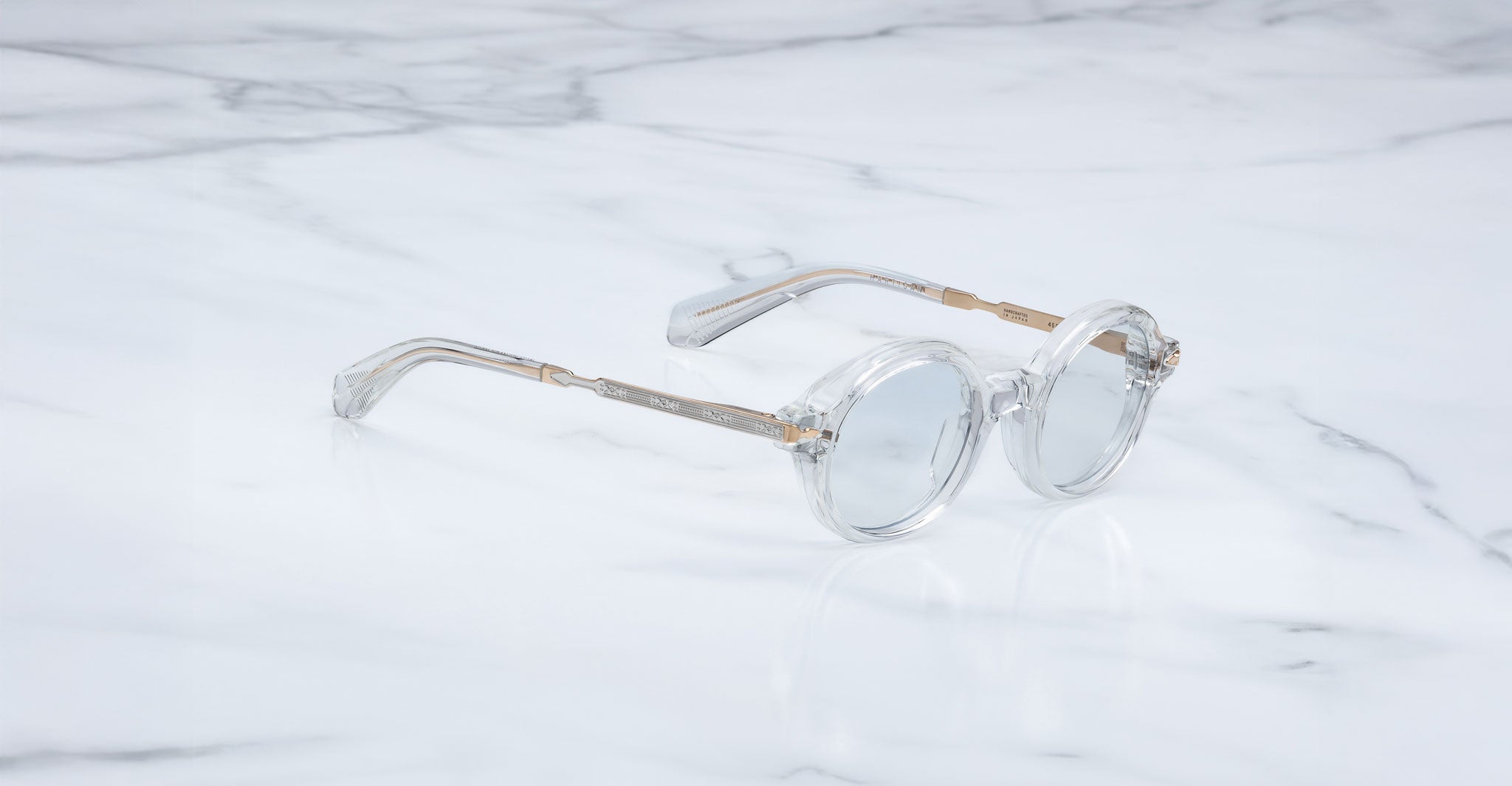 Jacques Marie Mage Hartigan in Clear, round transparent acetate eyeglasses shown from a side angle, highlighting engraved metal core temples, keyhole bridge, and sculpted temple tips on a light marble surface.