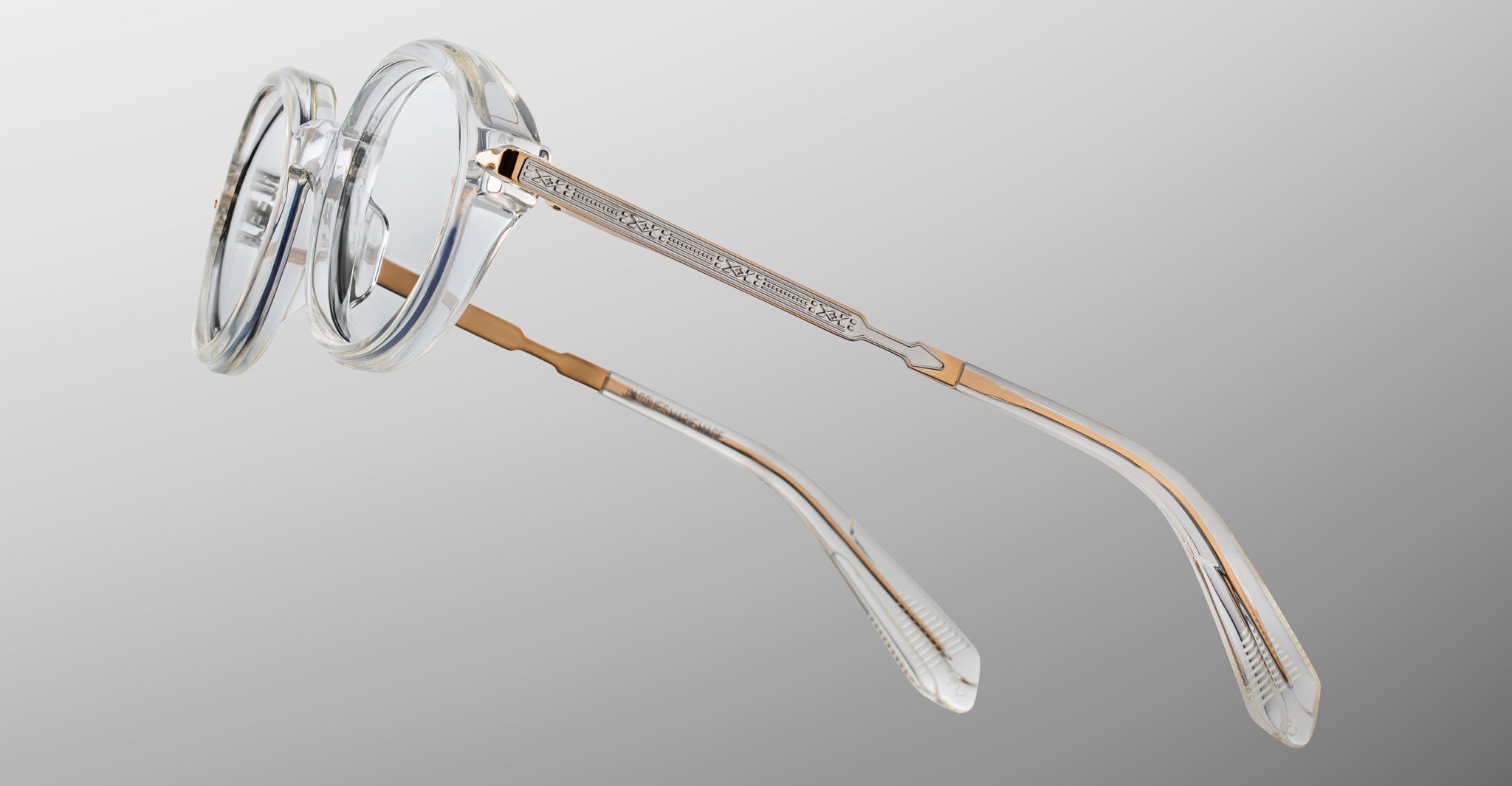 Jacques Marie Mage Hartigan in Clear, round transparent acetate eyeglasses shown from a rear side angle, highlighting engraved metal core temples, custom hinge details, and sculpted temple tips with clear demo lenses.