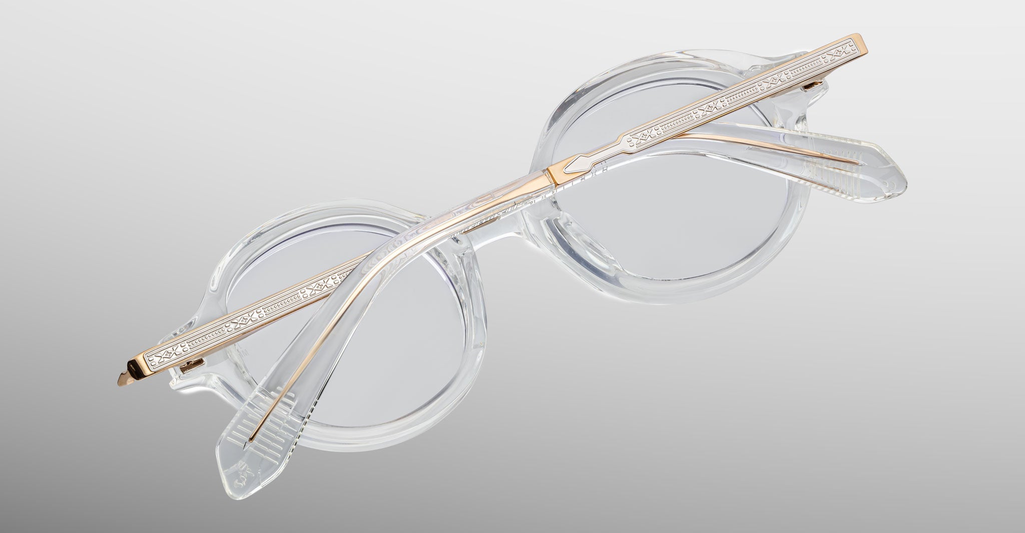 Jacques Marie Mage Hartigan in Clear, round transparent acetate eyeglasses shown from an angled top view, highlighting engraved metal core temples, keyhole bridge, and sculpted temple tips with clear demo lenses.