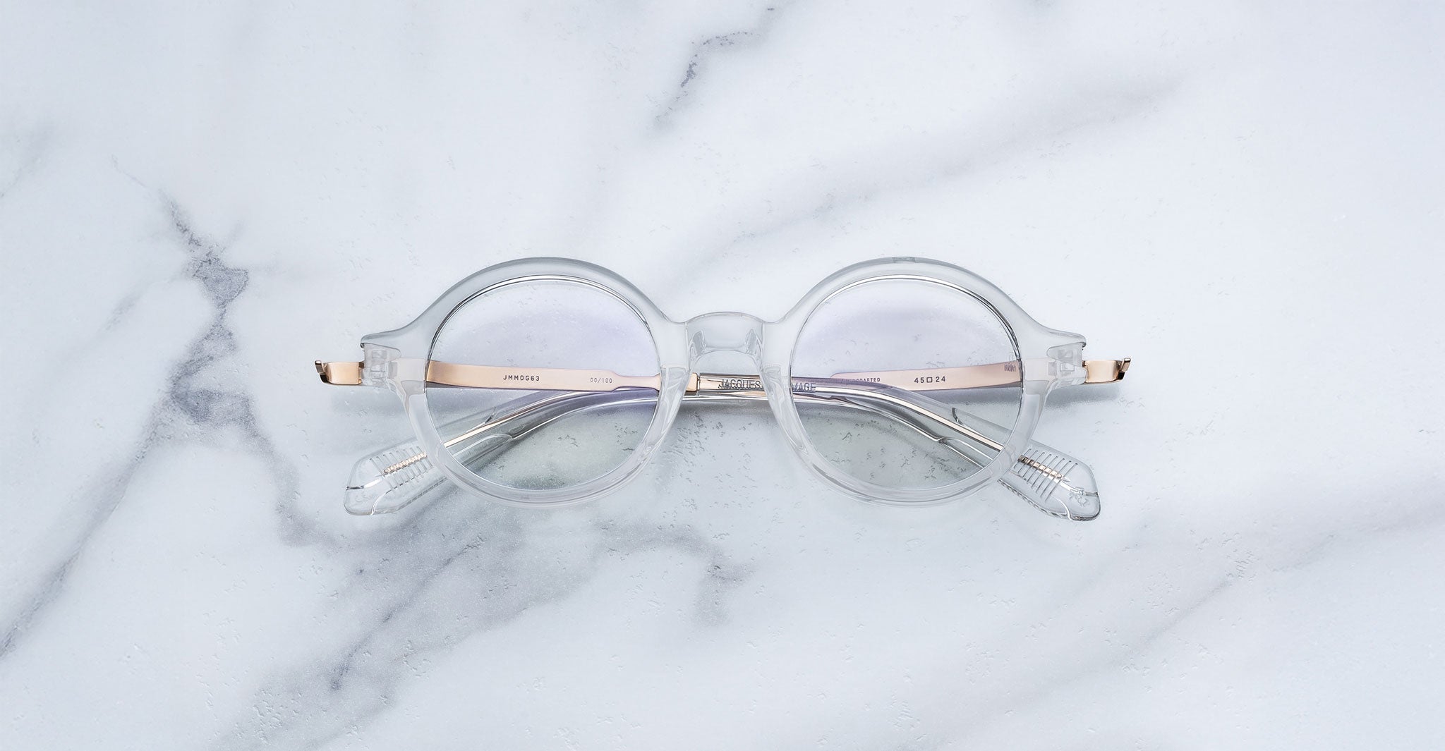 Jacques Marie Mage Hartigan in Clear, round transparent acetate eyeglasses with a keyhole bridge, exposed metal core temples, and subtle purple anti-reflective lens coating, shown front-facing on a light marble surface.