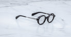 Jacques Marie Mage Hartigan in Phantom, round black acetate eyeglasses shown from a side angle, highlighting engraved metal core temples, keyhole bridge, and sculpted temple tips on a light marble surface.