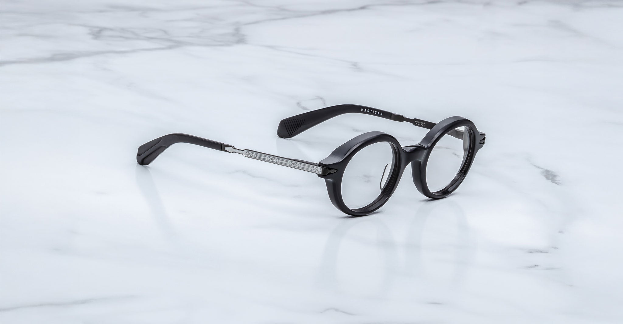 Jacques Marie Mage Hartigan in Phantom, round black acetate eyeglasses shown from a side angle, highlighting engraved metal core temples, keyhole bridge, and sculpted temple tips on a light marble surface.