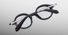Jacques Marie Mage Hartigan in Phantom, round black acetate eyeglasses shown from an angled top view, highlighting engraved metal core temples, keyhole bridge, and sculpted temple tips with clear demo lenses.