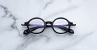 Jacques Marie Mage Hartigan in Phantom, round black acetate eyeglasses with a keyhole bridge, exposed metal core temples, and subtle purple anti-reflective lens coating, shown front-facing on a light marble surface.