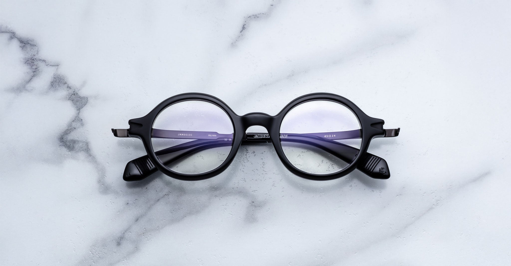 Jacques Marie Mage Hartigan in Phantom, round black acetate eyeglasses with a keyhole bridge, exposed metal core temples, and subtle purple anti-reflective lens coating, shown front-facing on a light marble surface.
