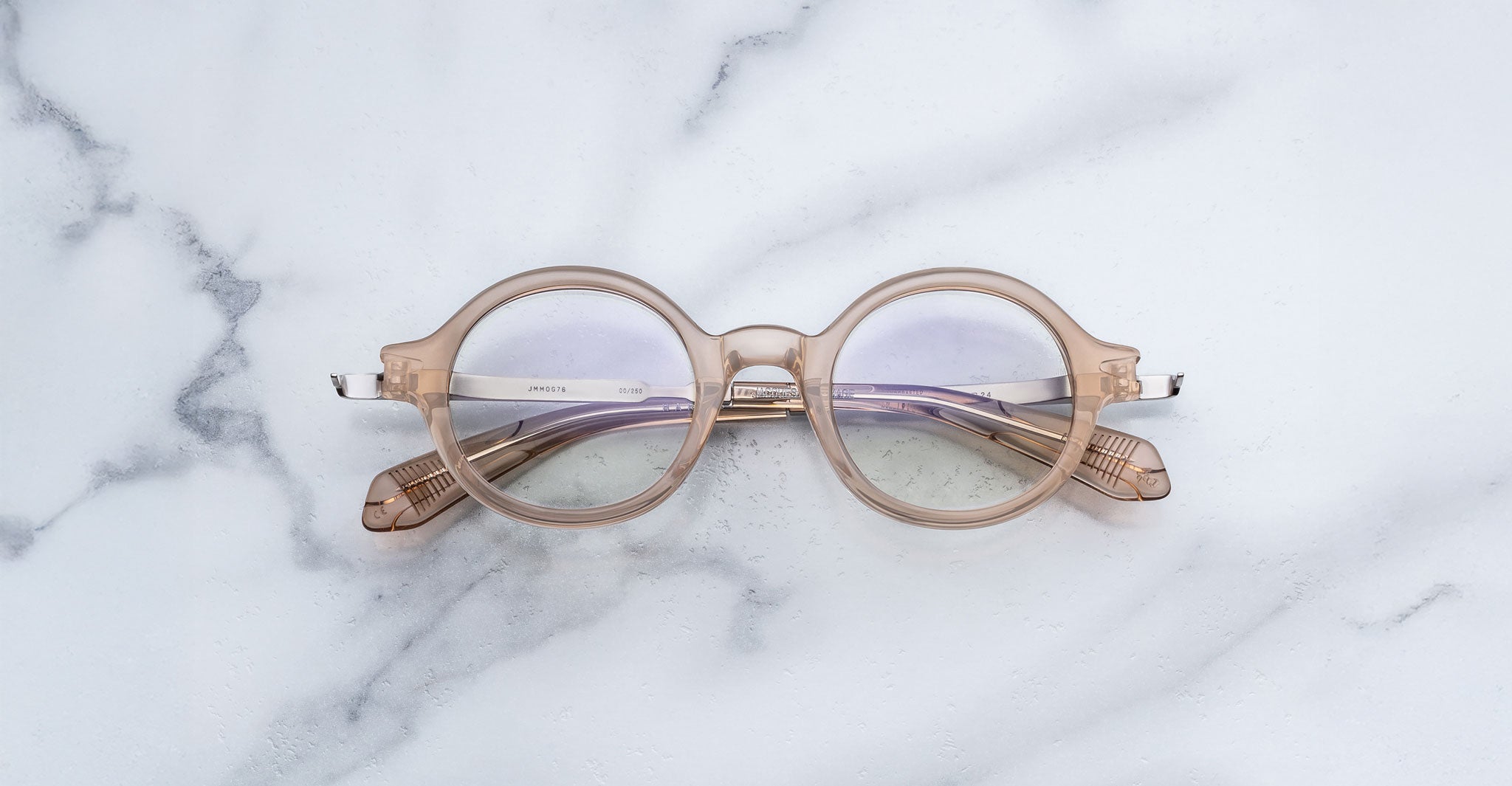 Jacques Marie Mage Hartigan in Sand, round translucent beige acetate eyeglasses with a keyhole bridge, exposed metal core temples, and subtle purple anti-reflective lens coating, shown front-facing on a light marble surface.