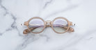 Jacques Marie Mage Hartigan in Sand, round translucent beige acetate eyeglasses with a keyhole bridge, exposed metal core temples, and subtle purple anti-reflective lens coating, shown front-facing on a light marble surface.