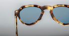 Close-up of Jacques Marie Mage Hatfield tortoiseshell eyeglass frames with round blue-tinted lenses against a plain, light gray background.