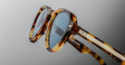 Close-up of Jacques Marie Mage Hatfield tortoise shell sunglasses featuring blue-tinted lenses, silver hinge details, and textured metal-accented temples set against a neutral gradient background.