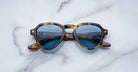 The Jacques Marie Mage Hatfield sunglasses, featuring brown tortoiseshell frames and blue-tinted lenses, are displayed on a white marble surface.