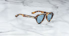 The Jacques Marie Mage Hatfield sunglasses, featuring a tortoiseshell pattern and blue tinted lenses, rest on a white marble surface.