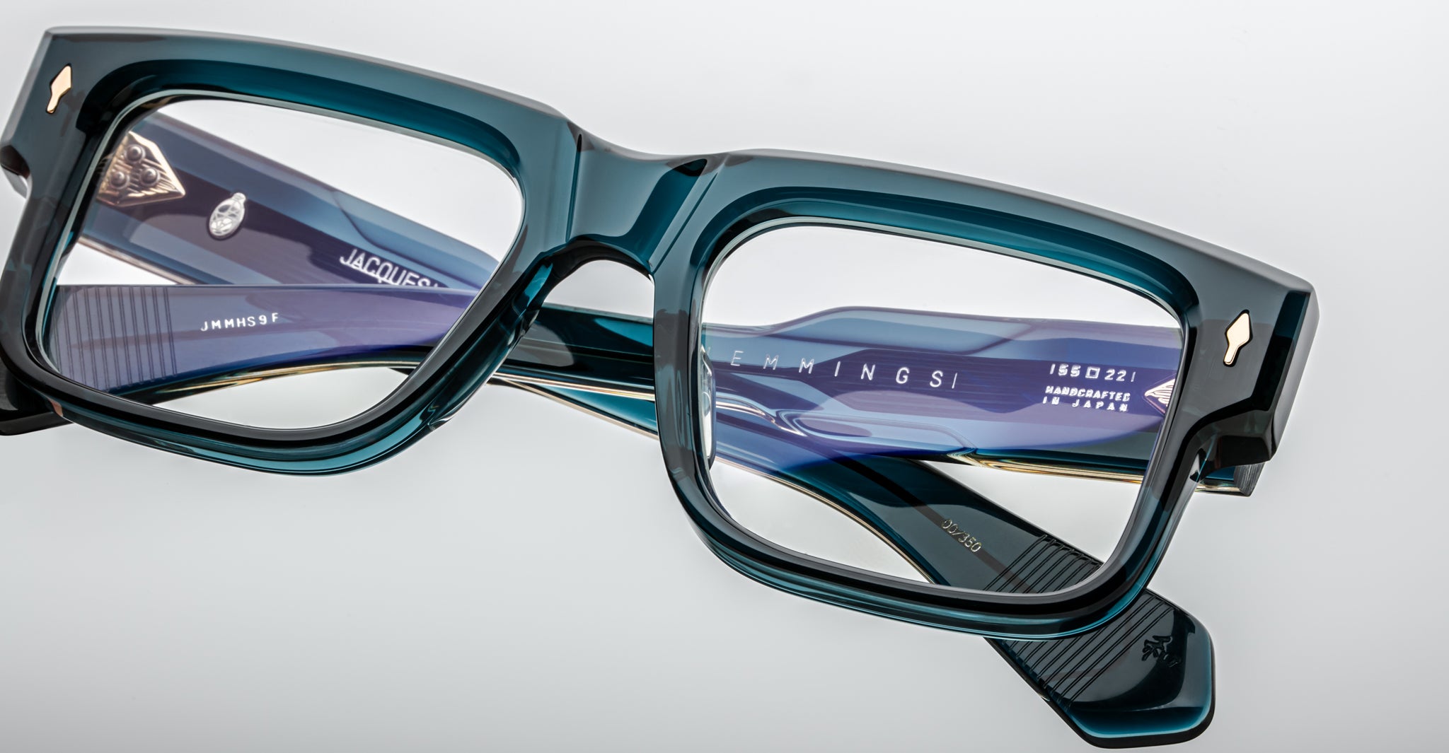 Close-up of Jacques Marie Mage Hemmings eyeglasses featuring stylish, thick-rimmed rectangular frames in teal blue, with detailed branding and inscriptions on the inner arms, resting on a glossy light surface.