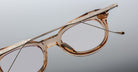 Close-up of Jacques Marie Mage Hisao eyeglasses—round, light brown frames with metallic engraved arms—displayed against a gradient gray background. One arm crosses over the other.