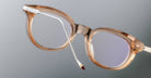 A close-up of the Jacques Marie Mage Hisao eyeglasses featuring light brown, translucent round frames, highlighting the end of one temple arm against a neutral grey background.