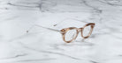 The Jacques Marie Mage Hisao eyeglasses, featuring a round shape in light brown with silver metal arms, rest on a white marble surface with gray veining.