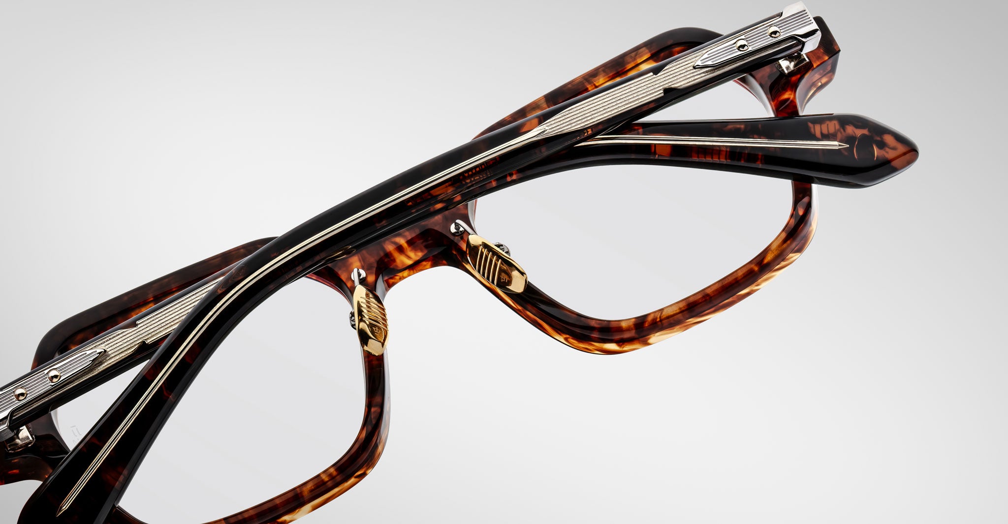 A close-up of two Jacques Marie Mage Holden eyeglasses with tortoiseshell frames and metallic hinges, stacked diagonally on a plain light gray background.