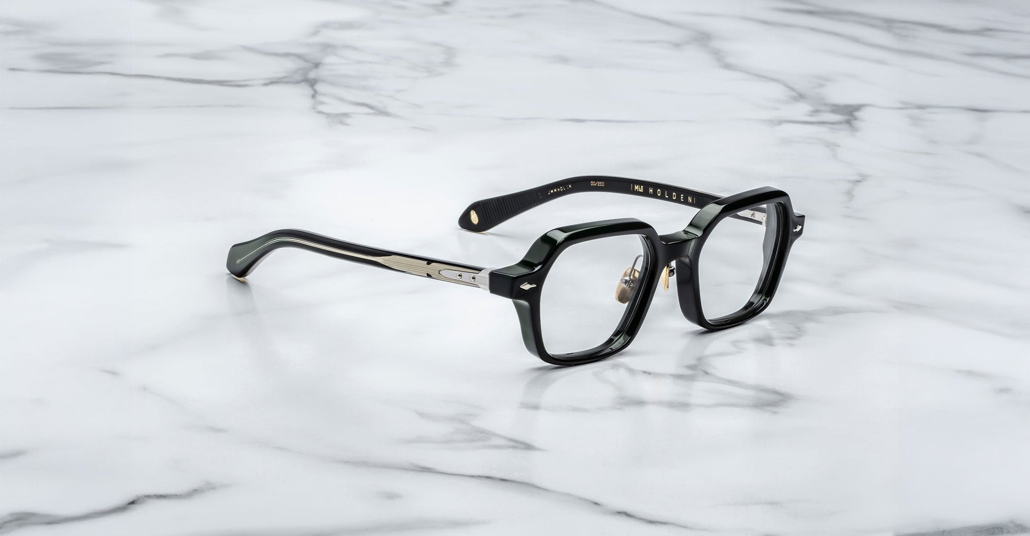 A pair of Jacques Marie Mage Holden black eyeglasses with thick rims and gold-accented temples rests on a white marble surface.