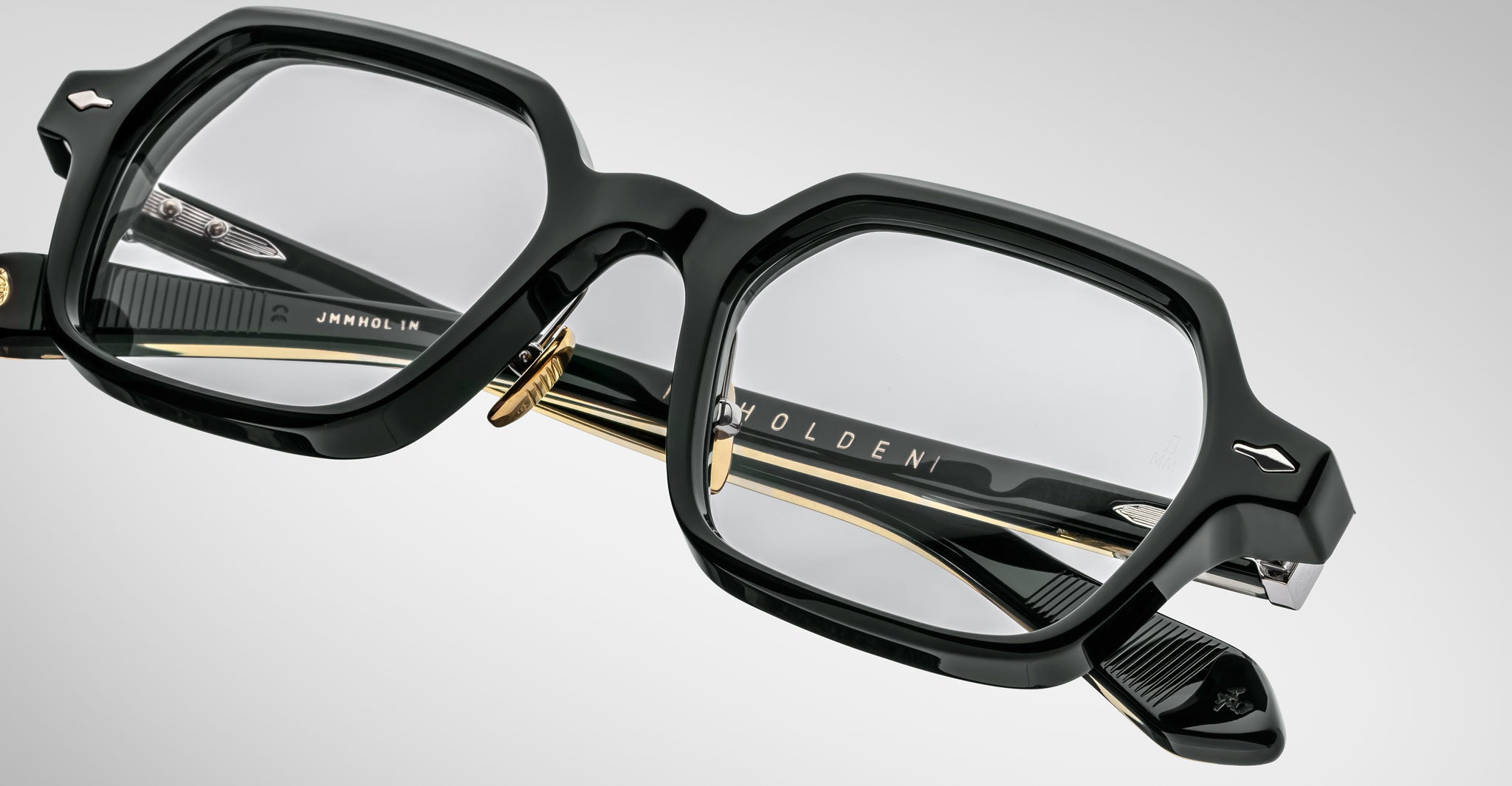 Close-up of Jacques Marie Mage Holden black, thick-framed eyeglasses with clear lenses and gold accents, showcasing detailed craftsmanship and branded arms against a soft light gray gradient background.