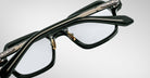 A close-up side view of the Jacques Marie Mage Holden eyeglasses in black and gold, highlighting their detailed metal arms and clear lenses against a light gray background.
