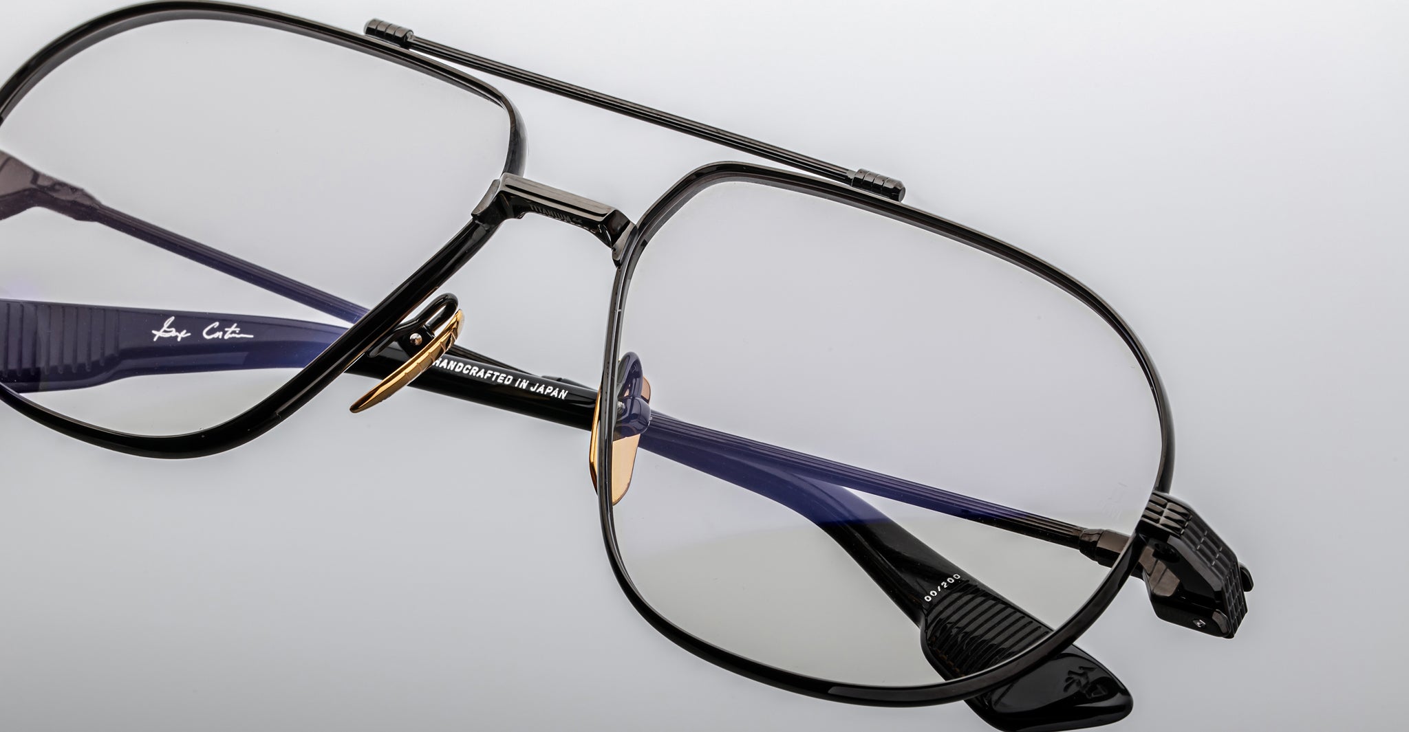 Close-up of Jacques Marie Mage Hollywood eyeglasses featuring stylish black rims, gold nose pads, and clear lenses on a light gray surface. One temple arm reads HANDCRAFTED IN JAPAN.