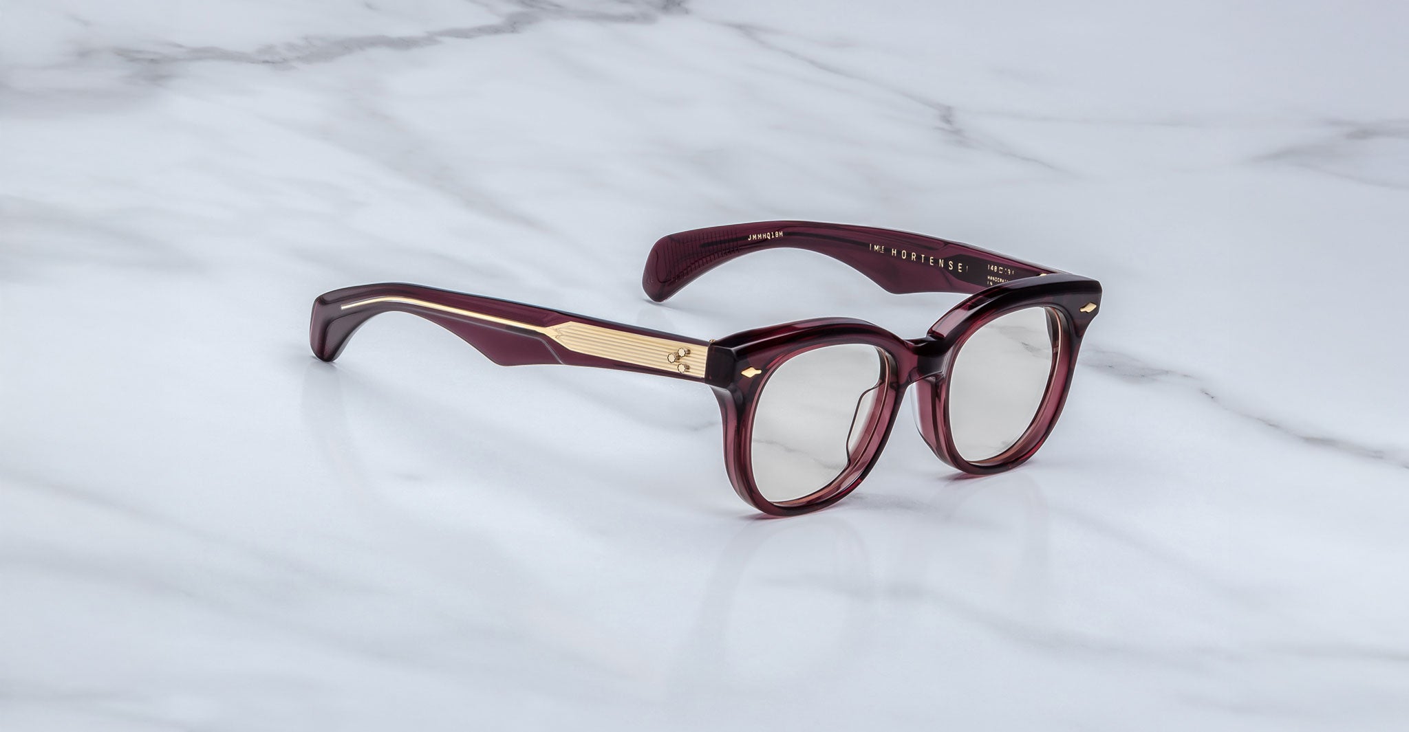 Jacques Marie Mage Hortense: Maroon round eyeglasses with clear lenses, gold metal temple accents, set against a marble-patterned background.