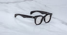 The Jacques Marie Mage Hortense eyeglasses feature black, thick round frames with subtle white speckles on the arms and silver accents by the hinges, displayed on a white marble surface.