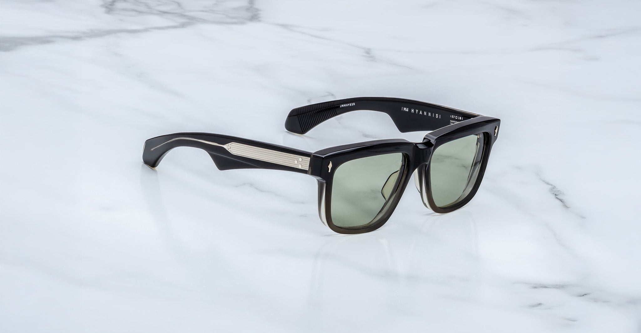 The Jacques Marie Mage Hyannis sunglasses feature thick, glossy rectangular frames with green-tinted lenses and metallic temple accents, combining modern style and limited-edition exclusivity on a white marble surface.