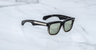 The Jacques Marie Mage Hyannis sunglasses feature thick, glossy rectangular frames with green-tinted lenses and metallic temple accents, combining modern style and limited-edition exclusivity on a white marble surface.