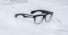 Jacques Marie Mage Hyannis rectangular eyeglass frames in a black-to-clear gradient with thick design and gold accents on the arms, shown on a white marble surface.