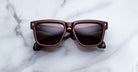 A pair of Jacques Marie Mage Hyannis limited-edition sunglasses with brown square frames and dark lenses rests on a white marble surface.