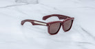 A pair of stylish Jacques Marie Mage Hyannis limited-edition maroon sunglasses with thick frames and dark lenses, displayed on white marble. The temples have metallic accents near the hinges.
