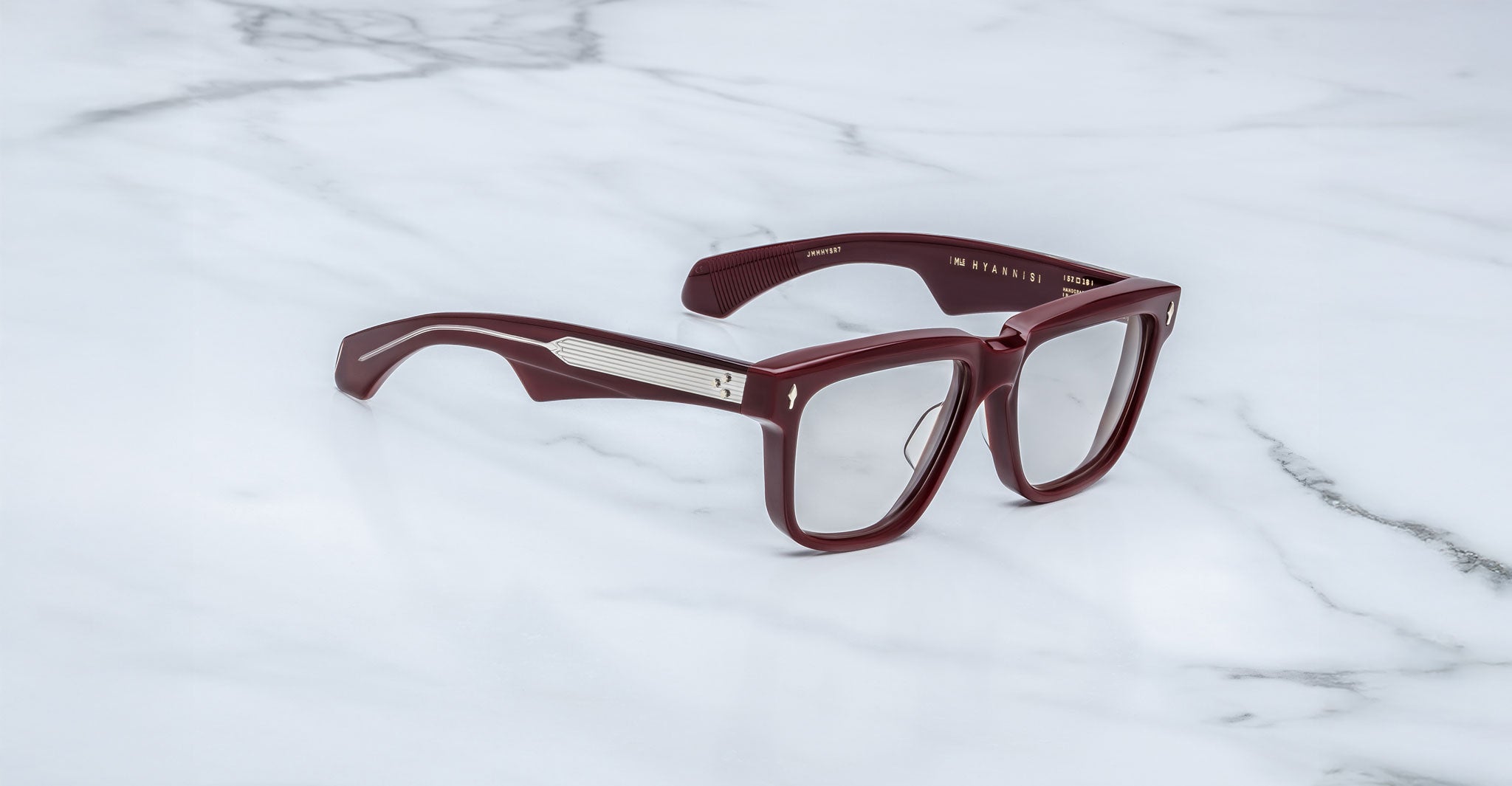 The Jacques Marie Mage Hyannis features thick, rectangular maroon frames with clear lenses and silver temple accents, shown on a white marble surface.