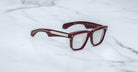 The Jacques Marie Mage Hyannis features thick, rectangular maroon frames with clear lenses and silver temple accents, shown on a white marble surface.