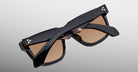 A pair of Jacques Marie Mage Hyannis limited-edition sunglasses with black rectangular frames and brown lenses, folded and set on a neutral gray background.
