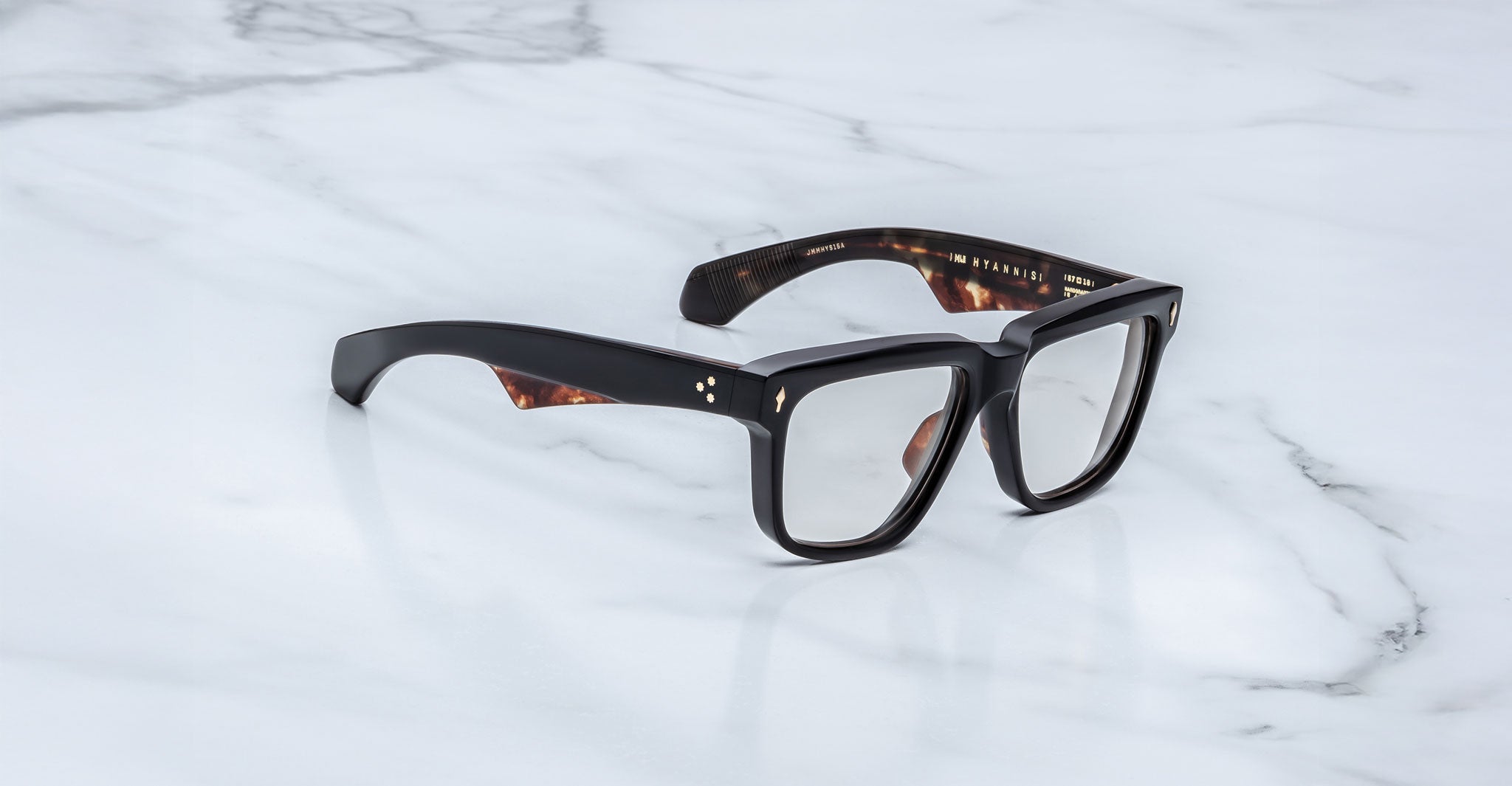 A pair of Jacques Marie Mage Hyannis rectangular eyeglass frames with thick black rims and inner tortoiseshell accents rests stylishly on a white marble surface.