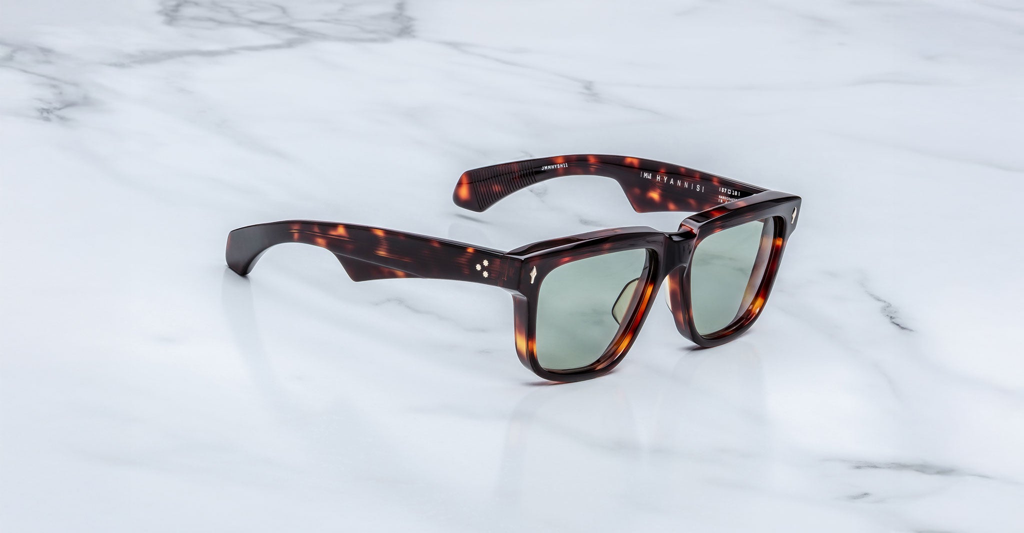The limited-edition Jacques Marie Mage Hyannis sunglasses feature thick, rectangular tortoiseshell frames and green tinted lenses, shown on a white marble surface with gray veining.