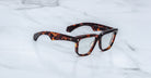 A pair of Jacques Marie Mage Hyannis eyeglasses with thick, rectangular tortoiseshell frames rests elegantly on a white marble surface.