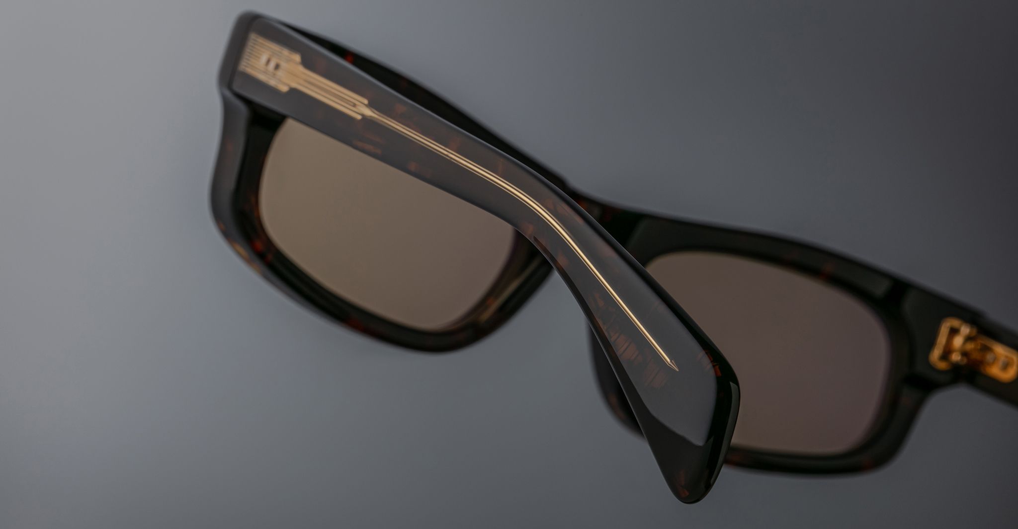 Close-up of Jacques Marie Mage Initials sunglasses—dark, rectangular frames with gold detailing on the arms—set against a softly lit gray background.