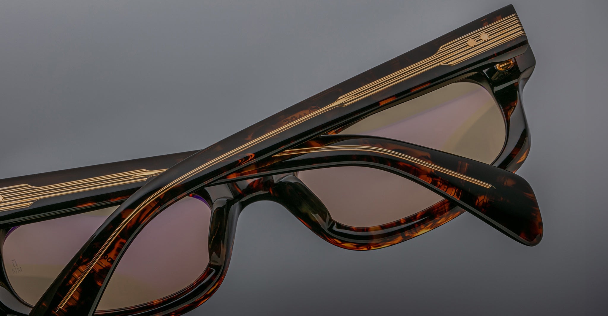 Close-up of two Jacques Marie Mage Initials tortoiseshell sunglasses with gold detailing, stacked against a plain gray background—highlighting the refined, limited-edition frames by Jacques Marie Mage.