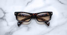 The Jacques Marie Mage Initials, a pair of brown tortoiseshell rectangular eyeglasses with thick frames, rests on white marble, highlighting the luxury of these limited-edition frames by Jacques Marie Mage.
