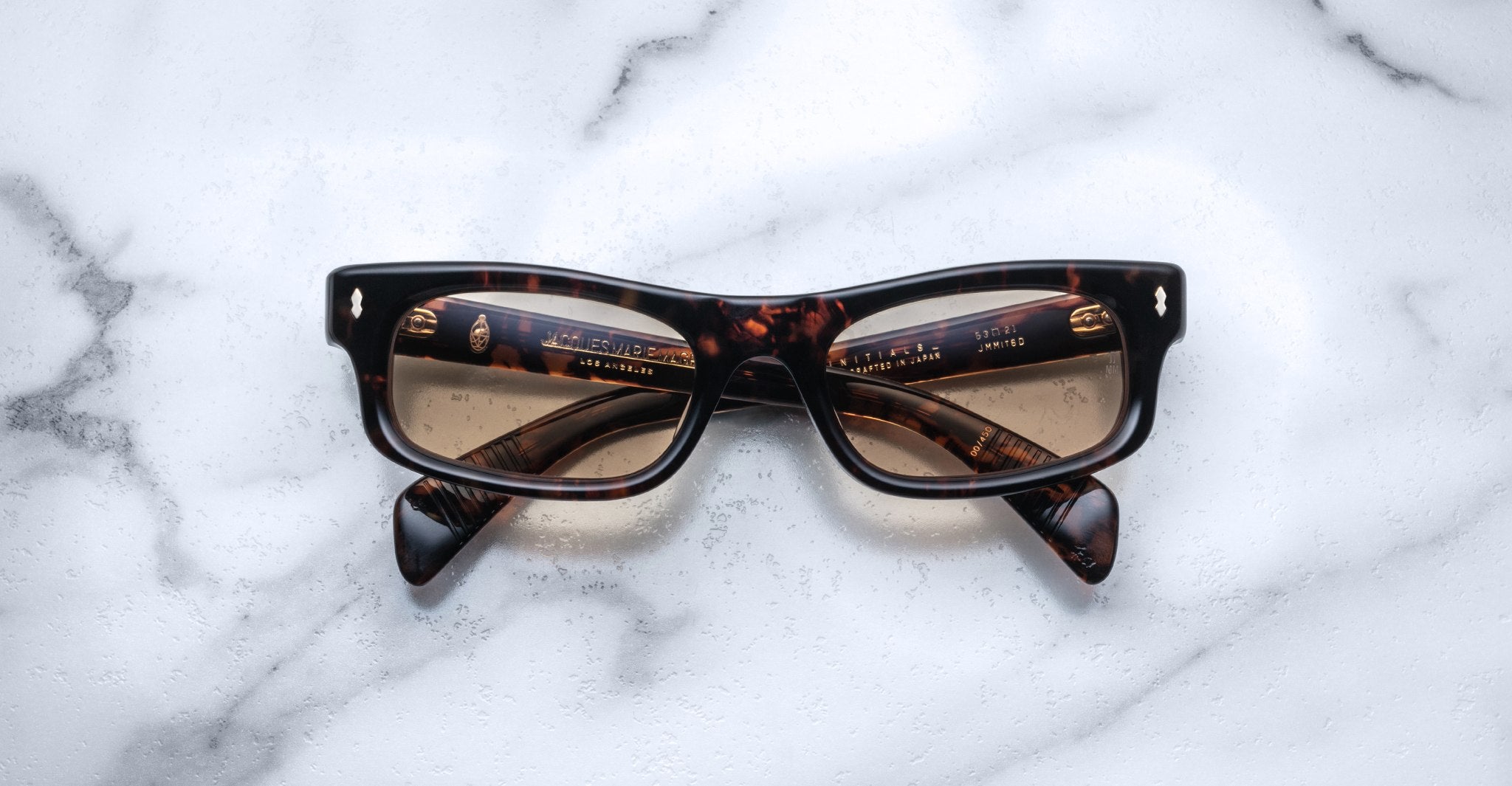 The Jacques Marie Mage Initials, a pair of brown tortoiseshell rectangular eyeglasses with thick frames, rests on white marble, highlighting the luxury of these limited-edition frames by Jacques Marie Mage.