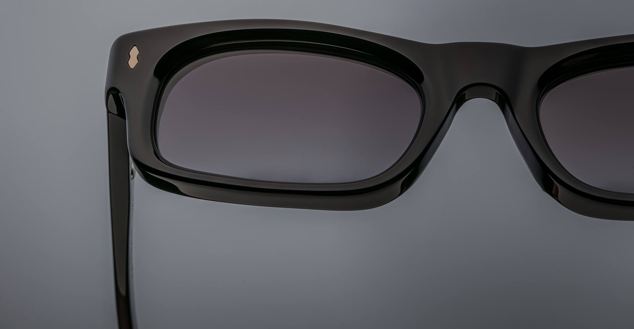 Close-up of Jacques Marie Mage Initials: limited-edition black rectangular micro-frame sunglasses with dark tinted lenses by Jacques Marie Mage, shown against a plain grey background.