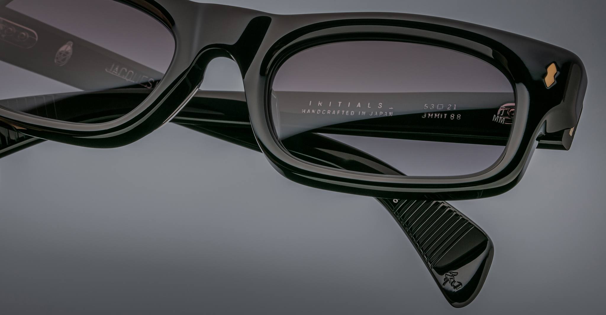 Close-up of Jacques Marie Mage Initials: black rectangular micro-frame sunglasses with gray tinted lenses, showcasing engraved inner-arm text and gold logo accents.