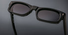 Close-up of Jacques Marie Mage Initials black rectangular sunglasses with dark lenses on a gray surface, highlighting the limited-edition frames.