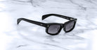 The Jacques Marie Mage Initials black rectangular micro-frame sunglasses with dark lenses rest on a white marble surface.