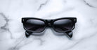 The Jacques Marie Mage Initials sunglasses feature a sleek black micro-frame and dark lenses, showcased on white marble. These limited-edition Jacques Marie Mage frames exude modern sophistication.