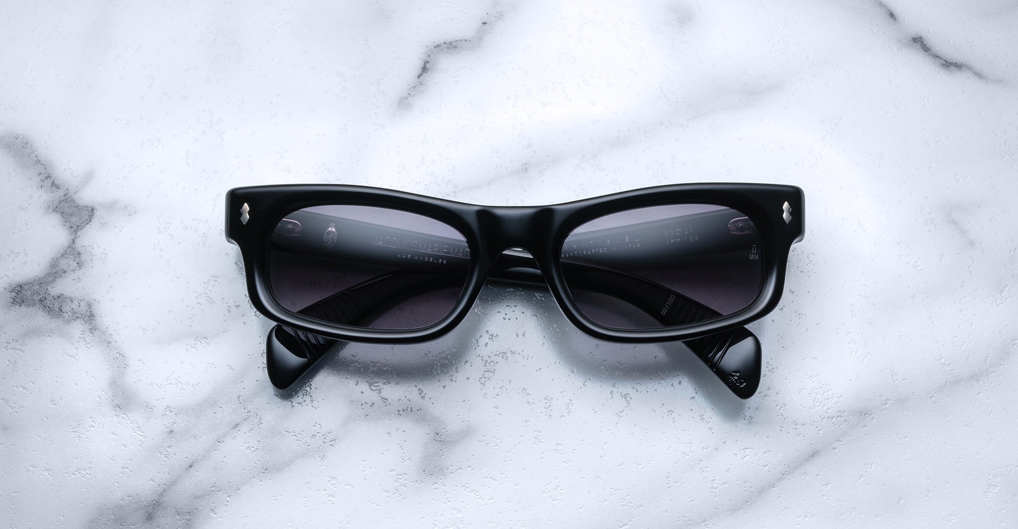 The Jacques Marie Mage Initials sunglasses feature a sleek black micro-frame and dark lenses, showcased on white marble. These limited-edition Jacques Marie Mage frames exude modern sophistication.